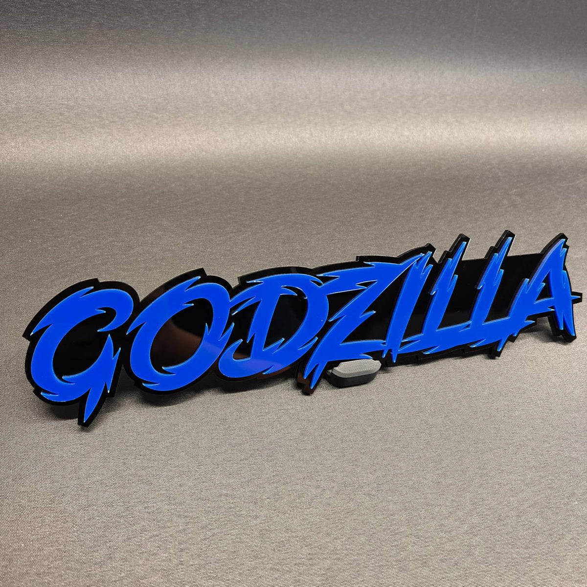 Godzilla auto badge with blue text and black backing for vehicle exterior