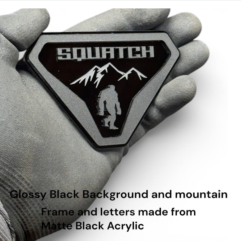 A hand wearing a gray work glove holds a glossy black triangular patch with a matte black acrylic frame. The patch features the word 'SQUATCH' in white above a white mountain and Bigfoot silhouette.