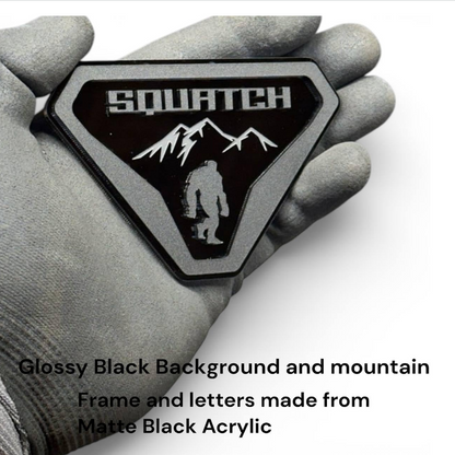 A hand wearing a gray work glove holds a glossy black triangular patch with a matte black acrylic frame. The patch features the word 'SQUATCH' in white above a white mountain and Bigfoot silhouette.