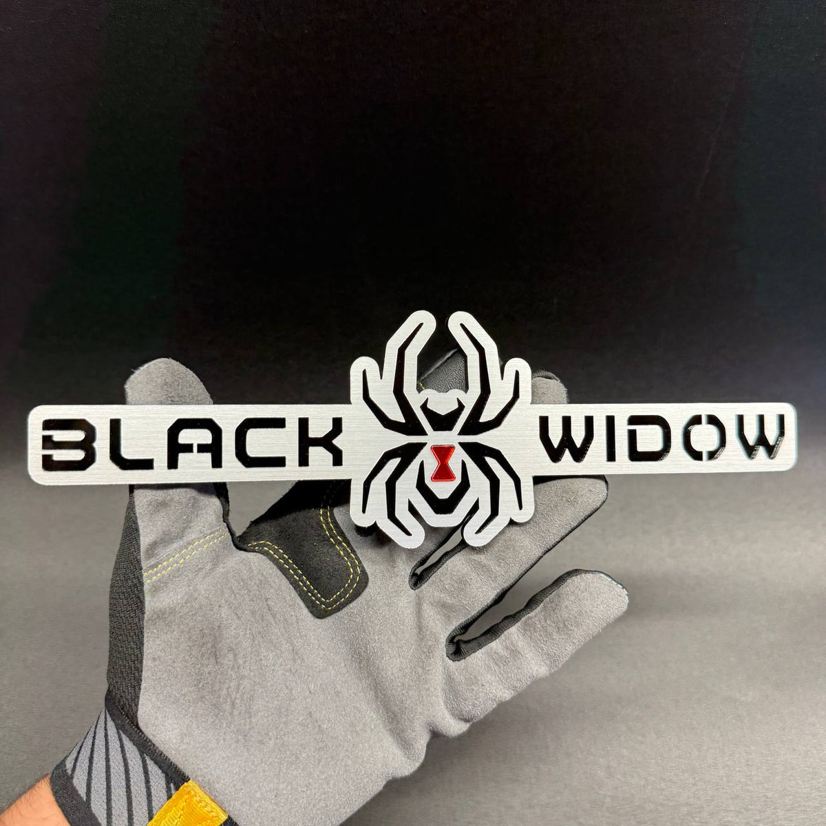 Black Widow spider car badge with black lettering, silver brushed background and red hourglass spider emblem.