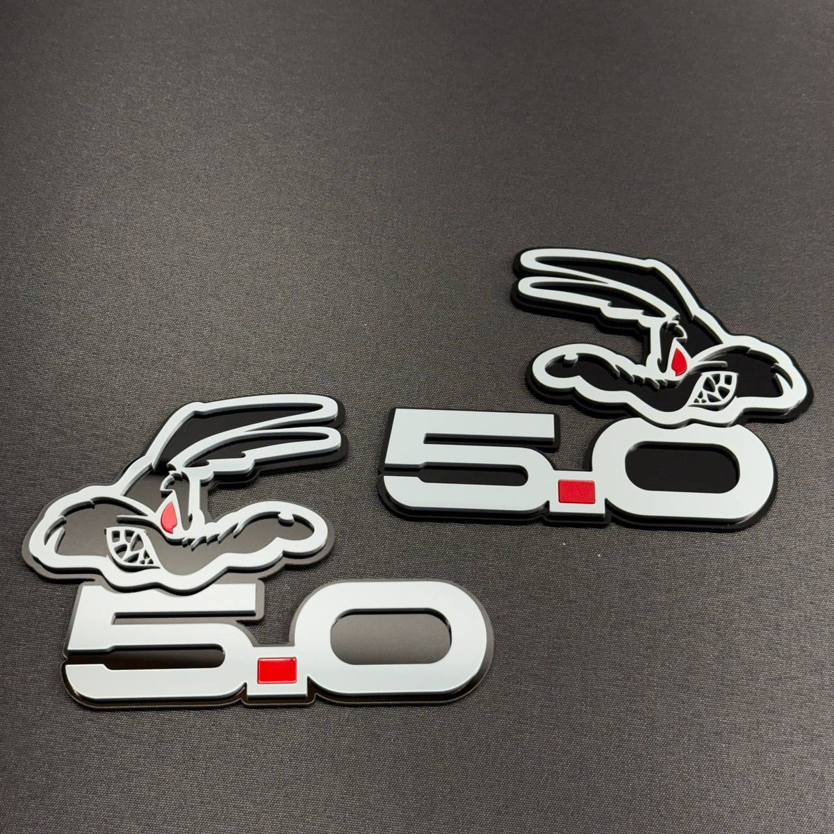12. Two Coyote 5.0 gray car badges displayed on textured fabric, showing left and right oriented cartoon coyote designs with red eyes.
