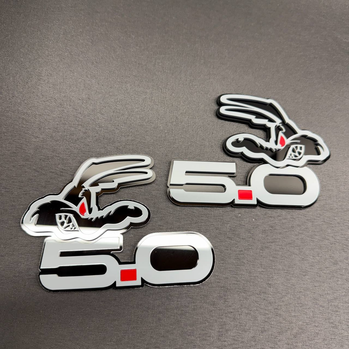 Ford Mustang 5.0 Coyote engine badge with gray finish, red square detail and black coyote character graphic.