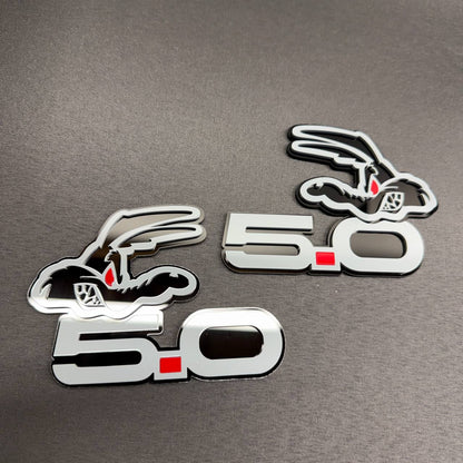 Ford Mustang 5.0 Coyote engine badge with gray finish, red square detail and black coyote character graphic.