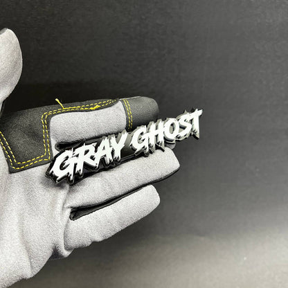 Gray Ghost Car Badge Fender Emblem shown on glove with white top and black backing
