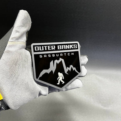 Black and silver Outer Banks Sasquatch car badge with mountain and Bigfoot design